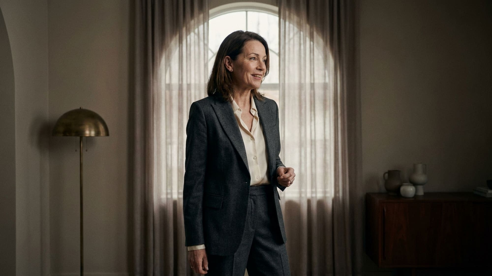 A woman in a tailored charcoal flannel suit standing in a spare, light-filled room
