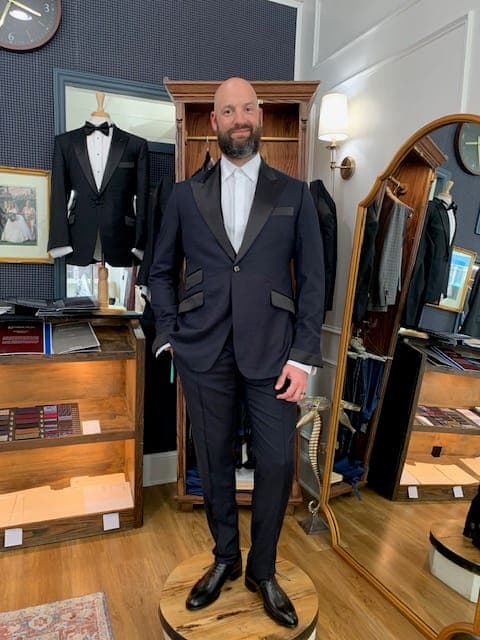 A client in a dinner jacket before the fitting-room mirror, tuxedos displayed behind